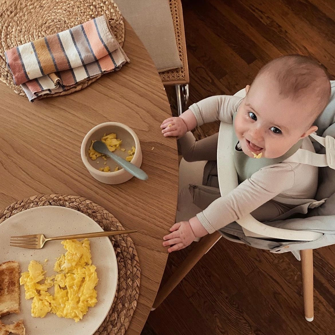 baby on a high chair smiling with her food in the cream aplainr silicone bowl on the table